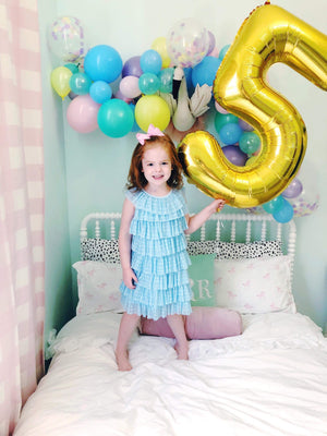 A little girl smiling and bounding on a white bed holding a gold number five balloon.