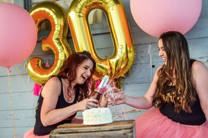 Two women laughing and saying cheers with gold 30 balloons floating behind them.