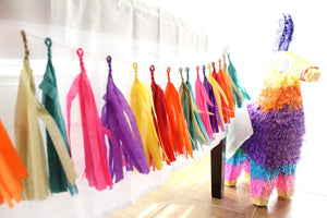 A fiesta themed llama pinata sits beside a tissue tassel garland strung with hot pink, purple, yellow, red, orange, gold and teal tissue tassels.
