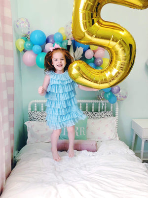 A jumbo gold number 5 balloon being held by a little girl jumping on her bed.