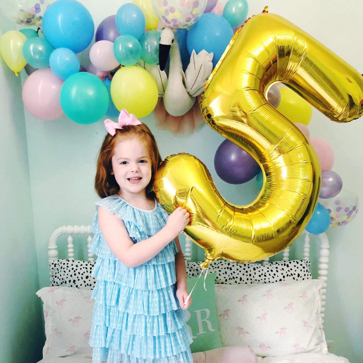 A little girl smiling and bounding on a white bed holding a gold number five balloon.