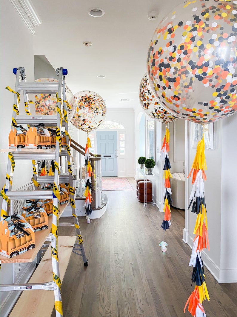 Three jumbo clear latex balloons filled with yellow, orange, black and white confetti with matching tissue tassel tails hanging from them float down a long hallway. To the left of the hallway is a shelve made of two ladders and wooden boards.