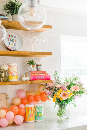 A mini balloon garland rests on top of a counter top underneath floating wooden shelves with a bouquet of fresh flowers sitting next to it. The balloon garland is made up of pink, coral, blush, and rose colored balloons.