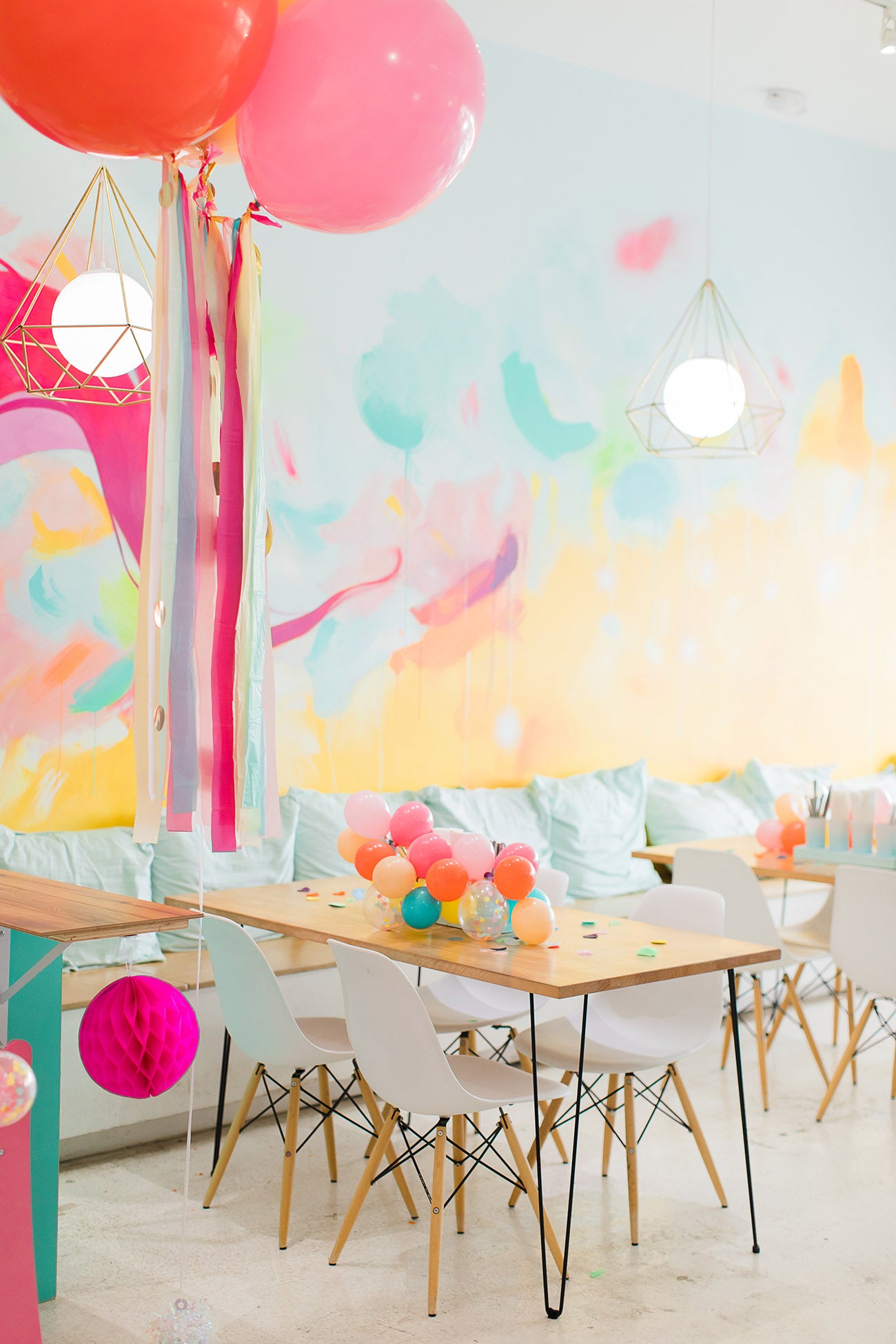 A very colorful setup shows a mini balloon garland sitting on a wooden table with jumbo red and pink helium inflated balloons floating beside it. The garland is made up of Caribbean blue, wildberry, coral, yellow, blush, and blush balloons.
