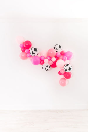A pink and cow print balloon garland hanging on a white wall.