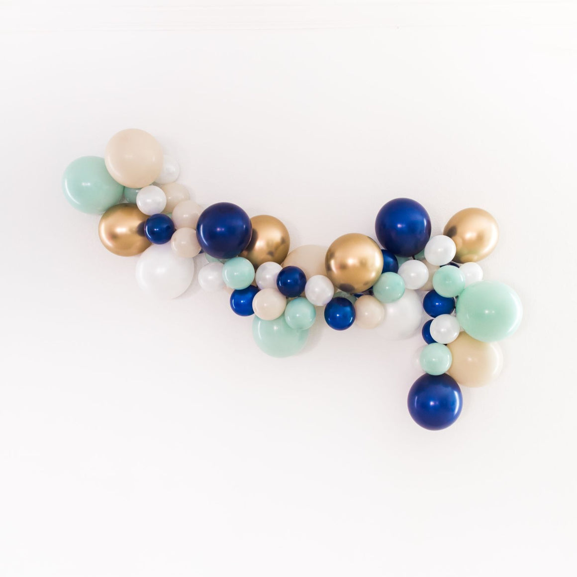 A nautical themed balloon garland is draped across a white wall. The garland consists of various 5 inch and 11 inch balloons in the colors of pearl white, tan, jade, navy, and chrome gold.