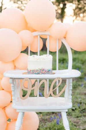 A white cake with small multicolored sprinkles on the bottom sits on the highchair with the wooden cursive one banner hanging.