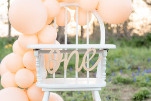 A white cake with small multicolored sprinkles on the bottom sits on the highchair with the wooden cursive one banner hanging.