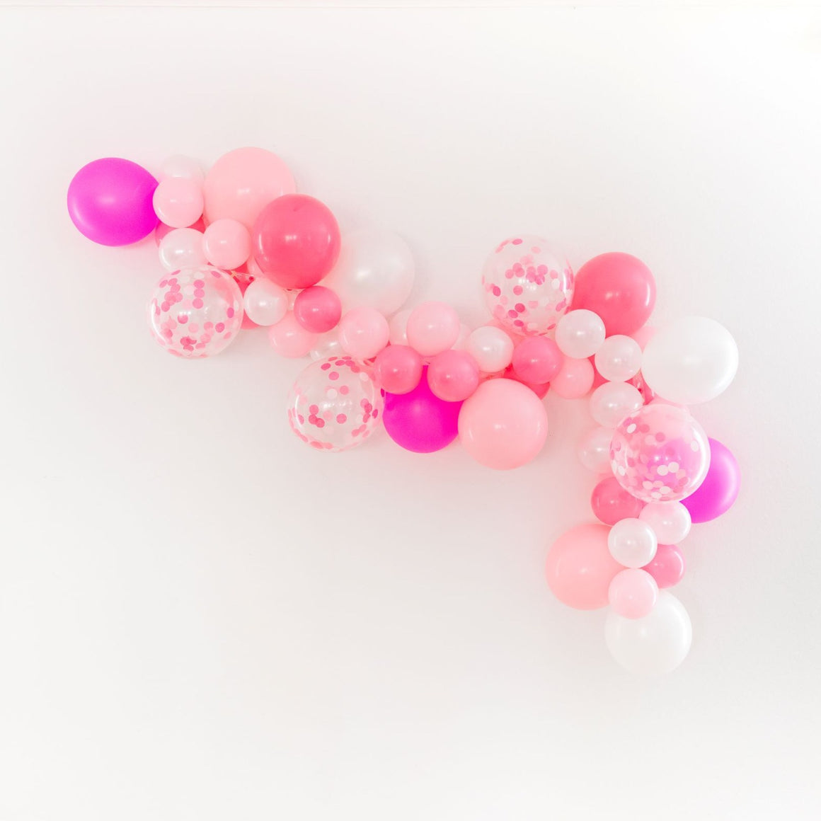 A side view of a pretty valentines themed garland is draped across a white wall. The garland is made of pink, rose, pearl white, neon magenta, and matching confetti balloons.