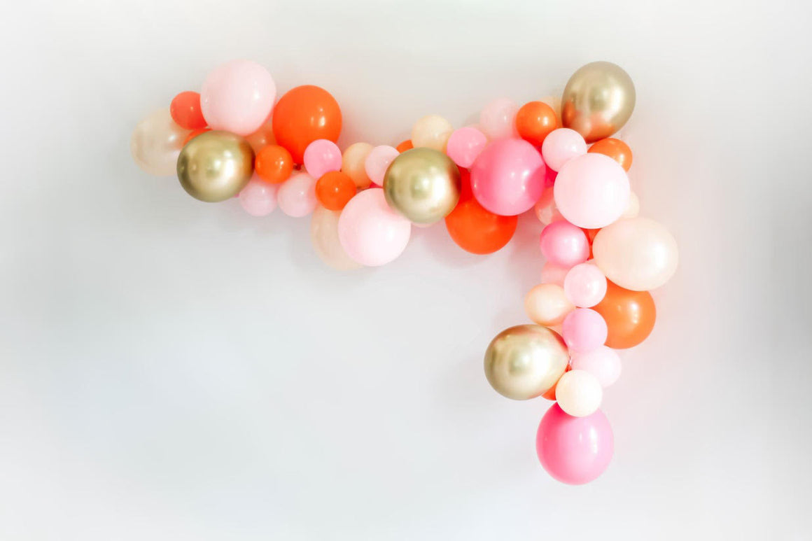 A pretty balloon garland is strung along a white wall. There is a mix of pink, rose, pearl peach, blush, orange, chrome gold balloons in both 5 inch and 11 inch sizes.