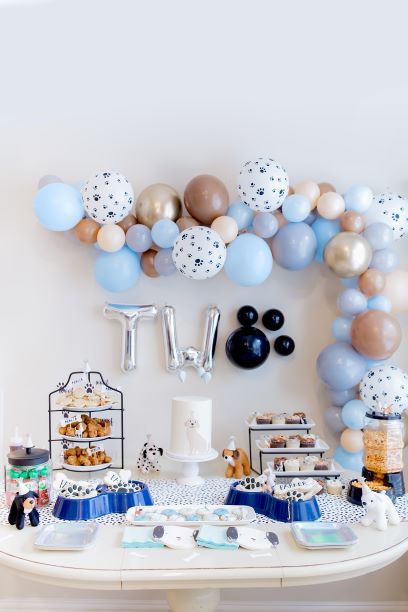 A puppy pawty set up celebrating a two year olds birthday. There are two silver balloons a T and a W followed by black balloons making a paw print. There is a balloon garland in the wall and a white table with cupcakes, cookies, and treats.