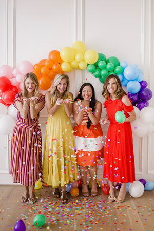 Four women blowing rainbow confetti from their hands standing in front of a balloon arch garland.
