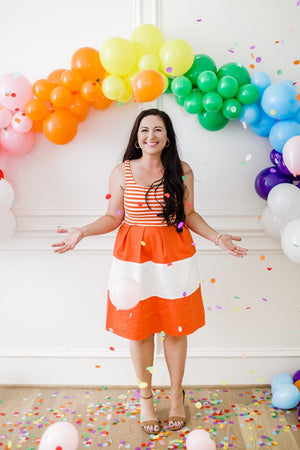 A woman in an orange and white dress is throwing rainbow colored confetti. There is a rainbow balloon garland behind her on a wall.