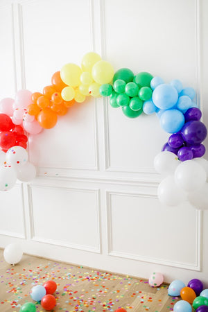 Rainbow balloon arch garland with white balloons at the ends hanging from a white wall. There are rainbow colored confetti and balloons on the floor.
