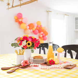 A bright fun balloon garland is draped across a white wall. The garland is made of a mix of 5 and 11 inch balloons in the following: orange, goldenrod, coral, blush, pink, and rose.