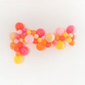 A bright fun balloon garland is draped across a white wall. The garland is made of a mix of 5 and 11 inch balloons in the following: orange, goldenrod, coral, blush, pink, and rose.