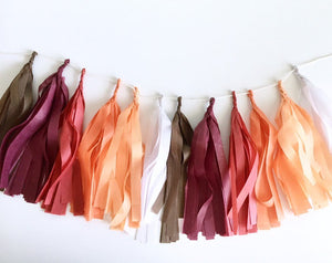An arial view of a brown, maroon, red, orange, peach, and white tissue tassel garland sitting on a white table.