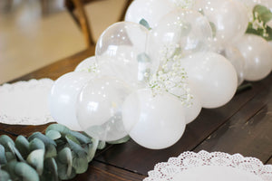 A soft 5 inch mini balloon garland made with clear, pearl white, and white balloons sits on a dark wooden table with a few rich colored florals scattered around.