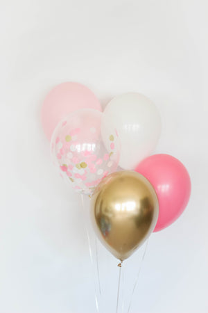 A five balloon bundle with one pink, one rose, one white, one chrome gold, and one confetti filled balloon in front of a white wall.