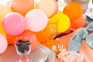 A 5 inch mini balloon garland with pink, rose, yellow, and orange balloons sits on a table behind a plate holding a clear glass cup filled with blackberries.