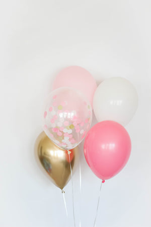 A balloon bundle with one pink, one rose, one white, one chrome gold, and one confetti filled balloon in front of a white wall.