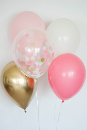 A girls balloon bundle with one pink, one rose, one white, one chrome gold, and one confetti filled balloon in front of a white wall.