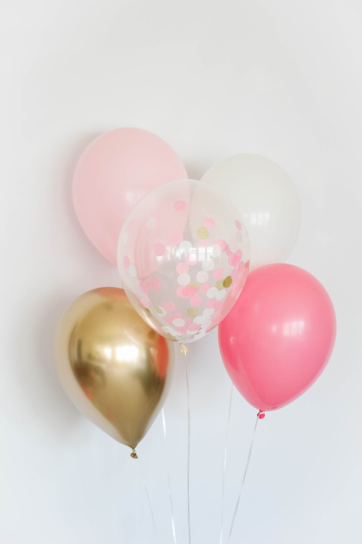 A five balloon bundle with one pink, one rose, one white, one chrome gold, and one confetti filled balloon in front of a white wall.