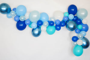Eight foot blue themed balloon garland on a white wall. The colors of the balloons are white, light blue, pearl light blue, blue, periwinkle, caribbean blue, and chrome blue.