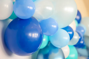 Close up of an eight foot blue themed balloon garland on a white wall. The colors of the balloons are white, light blue, pearl light blue, blue, periwinkle, caribbean blue, and chrome blue.