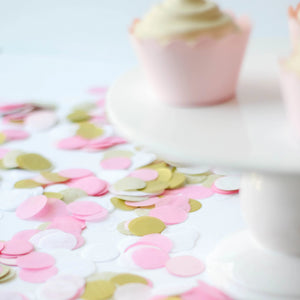 A pile of 1 inch handcut confetti circles in the colors pink, blush, white, and gold used in party celebrations, bachelorette, princess party, or girls birthday. A white cake stand with cupcakes sits on the right side of the photo.