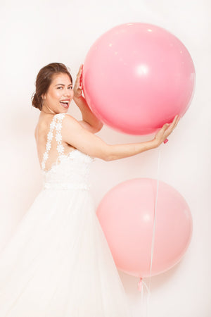 Girl smiling in a white dress holding a giant pink balloon and looking at the camera.